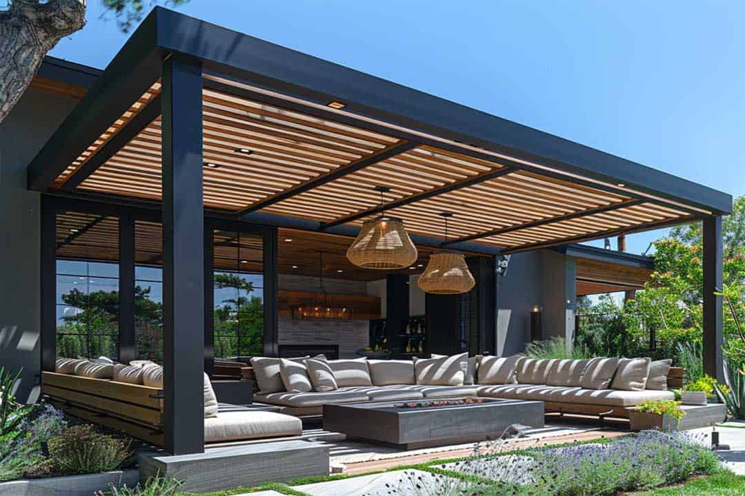 Attached aluminum patio cover with slatted wood roof, sectional seating, and fire table in a landscaped backyard.